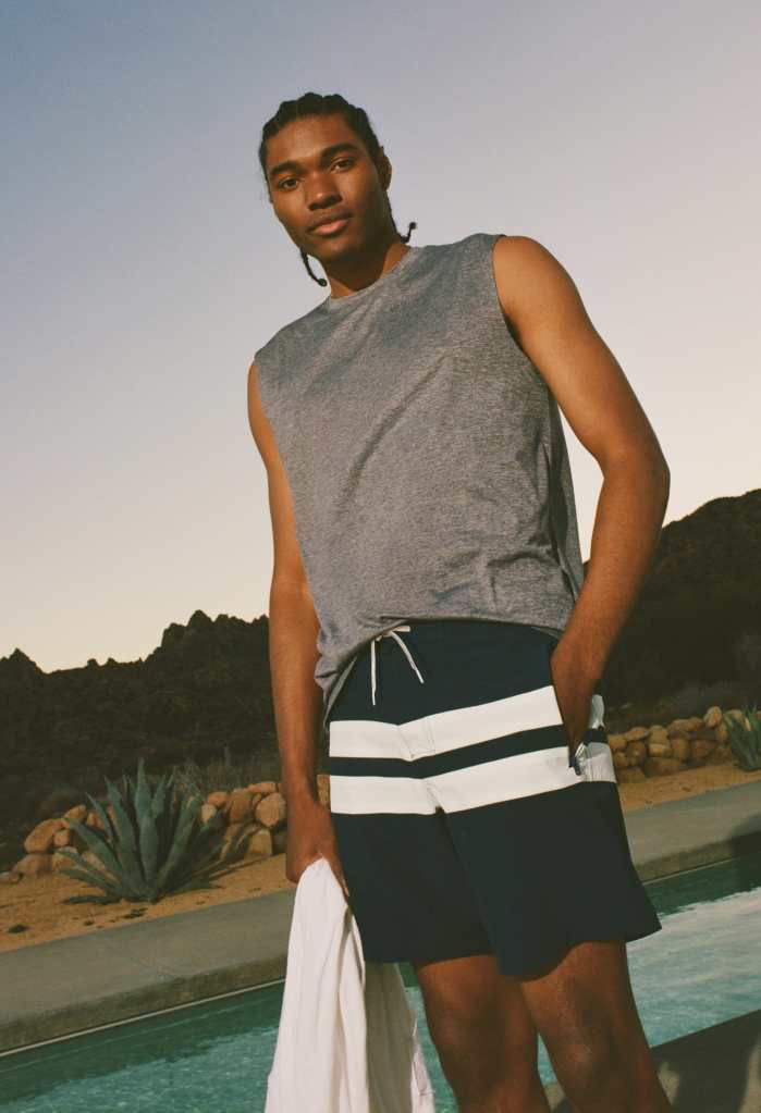 guy standing outside in front of a lake wearing striped blue swim shorts and a gray tank top from American Eagle