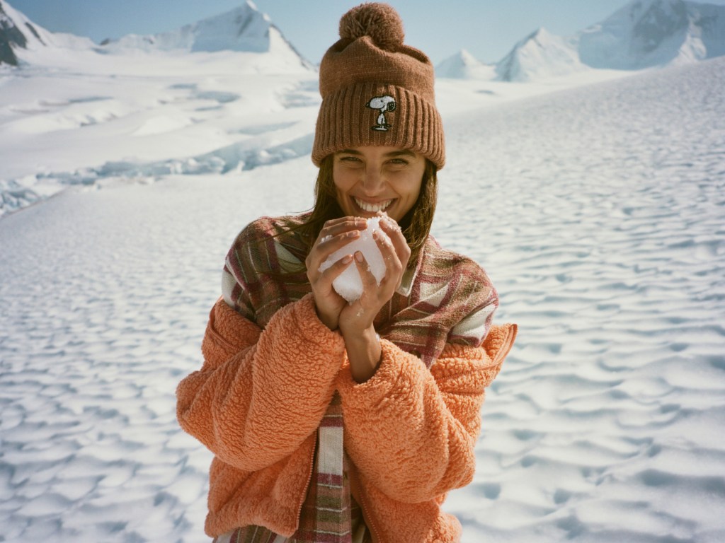 girl in a snowy landscape wearing an organge sherpa jacket, plaid shacket, and snoopy hat from American Eagle