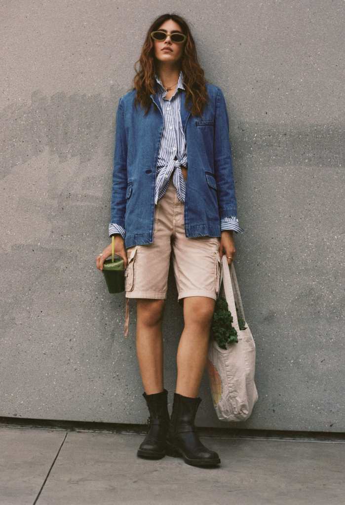 girl standing in front of a concrete wall wearing tan American Eagle shorts, a button-up shirt, and a denim jacket